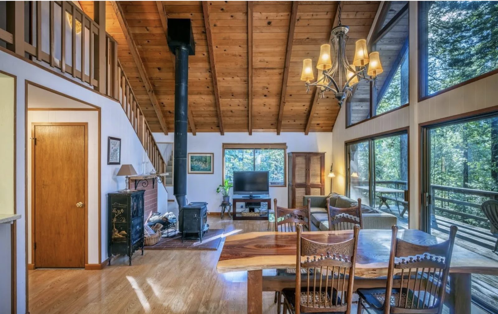 The living room before the remodel: knotty pine ceilings, a black wood stove and stovepipe on a brick hearth, dated furniture and laminate floors, redwoods visible through the windows.